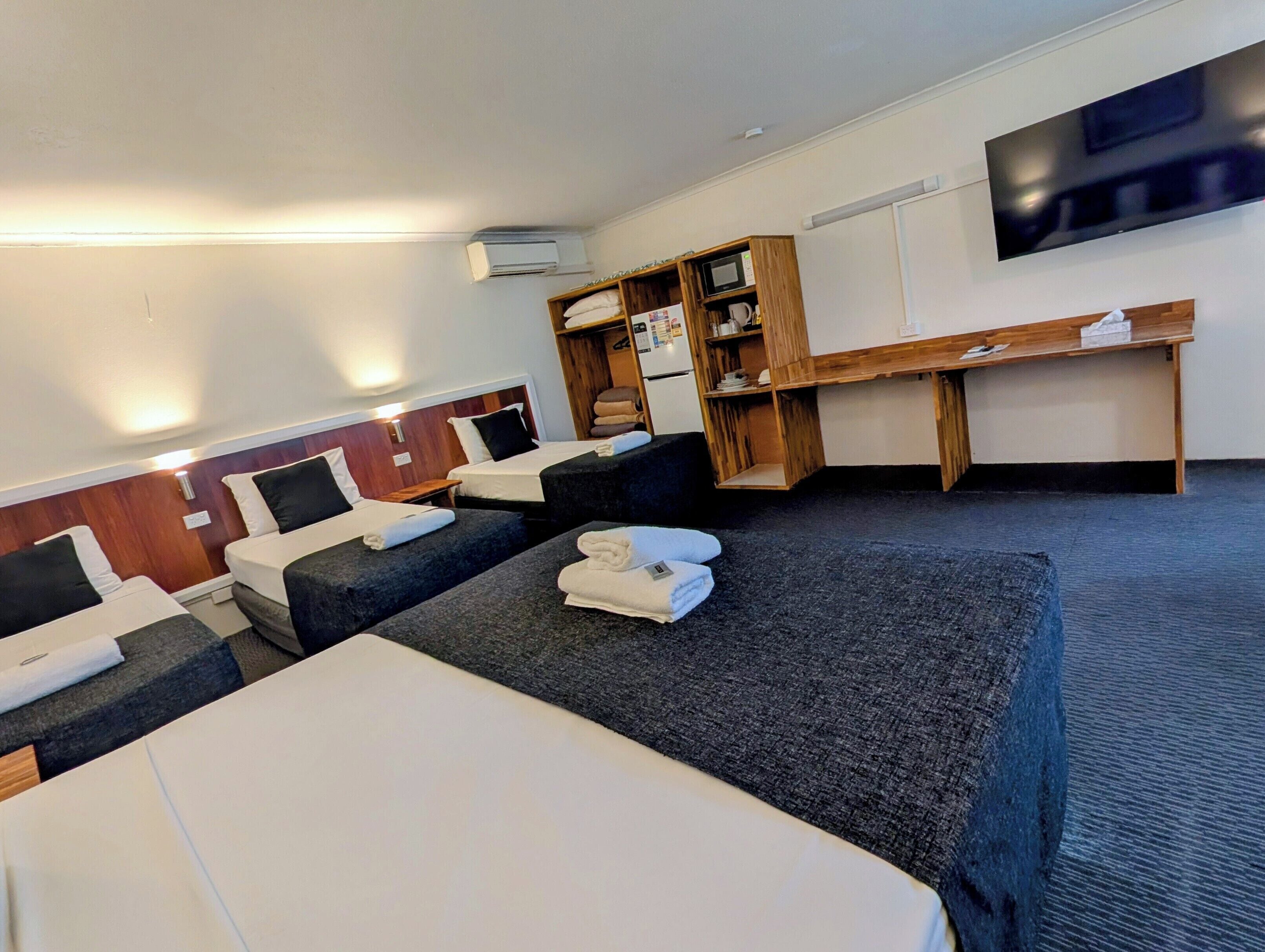 A hotel room with three beds, a flat-screen TV mounted on the wall, a wooden desk, and a built-in microwave.
