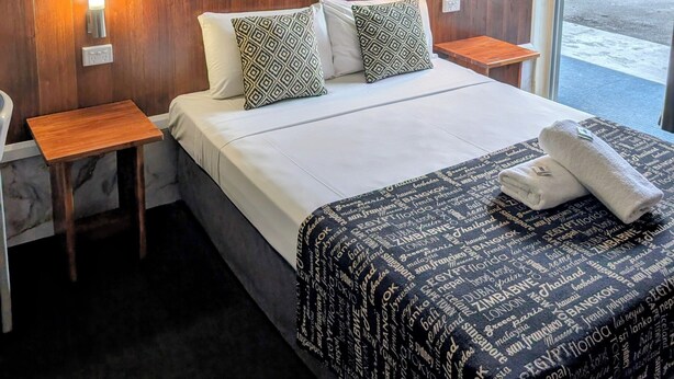A hotel room with a marble-patterned headboard, a bed with patterned pillows, a bedside table, and a window with a view of the parking area. - Hampton Villa Motel (Rockhampton)