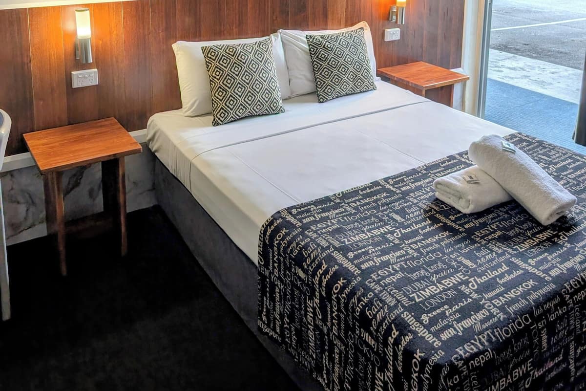 A hotel room with a marble-patterned headboard, a bed with patterned pillows, a bedside table, and a window with a view of the parking area.