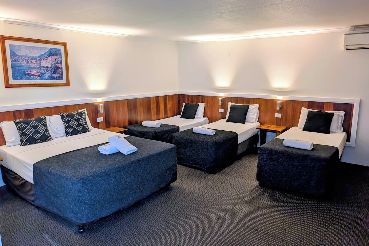 A hotel room with four beds arranged in a row, each with a pillow and a folded towel. There is a painting of a Mediterranean view on the wall.