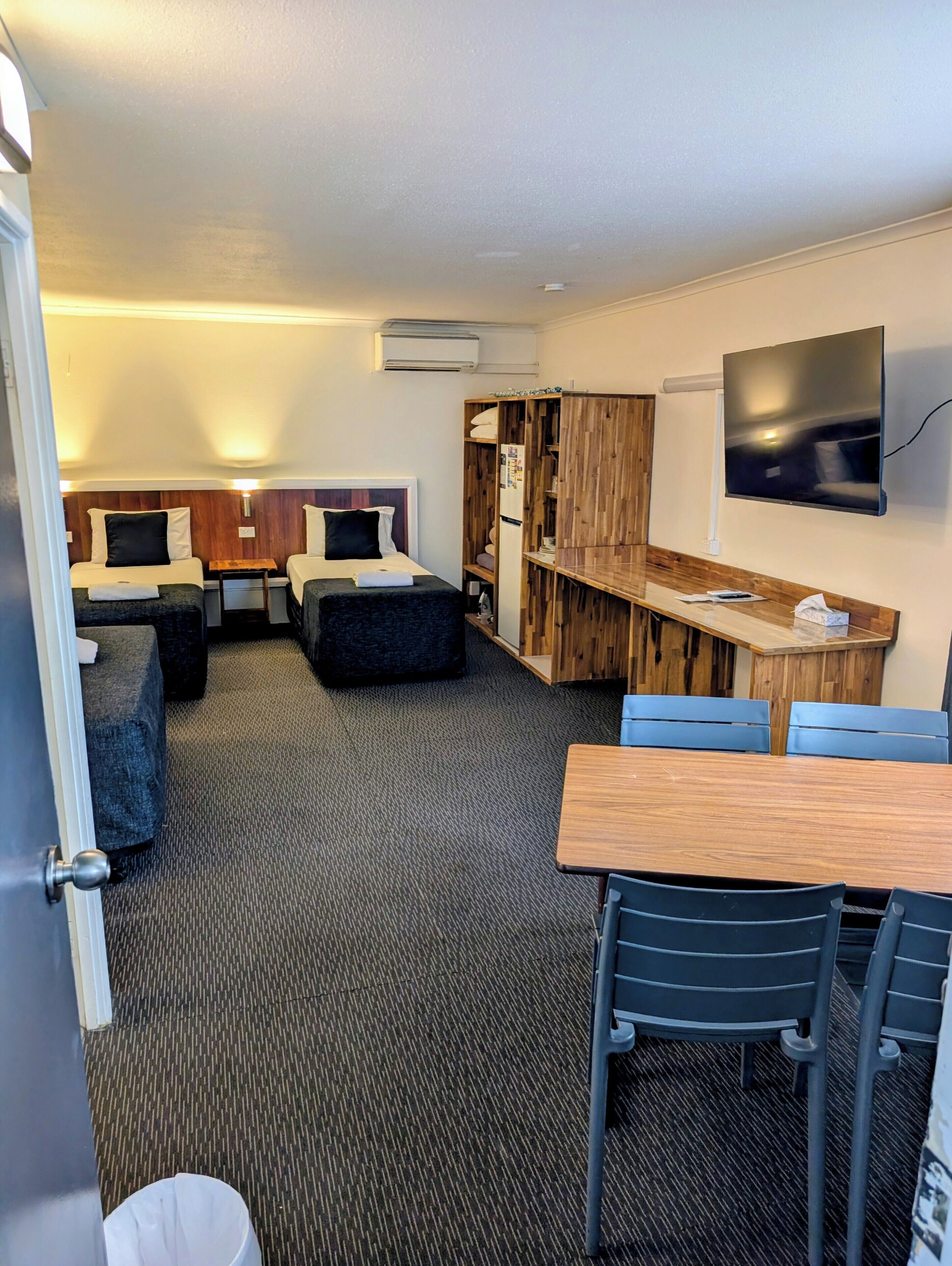 A compact hotel room with two single beds, a small table, a TV, and a kitchenette area.