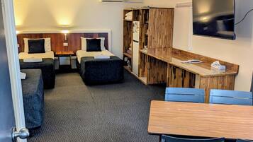 Quarto de hotel compacto com duas camas de solteiro, uma mesinha, televisão e área de cozinha compacta.