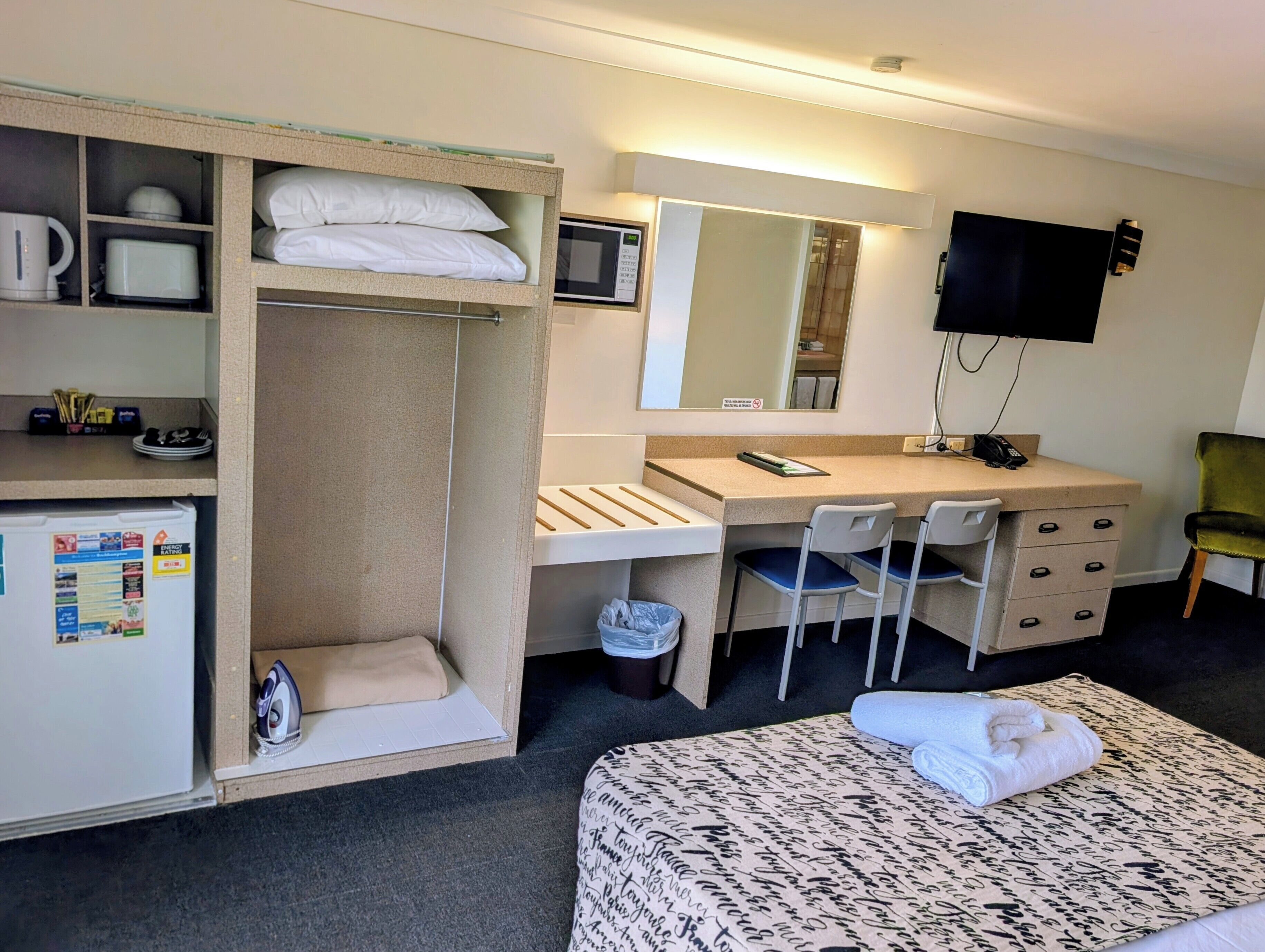 Habitación de hotel con cama, escritorio con dos sillas, microondas, refrigerador, armario, cesto de basura y televisor montado en la pared.