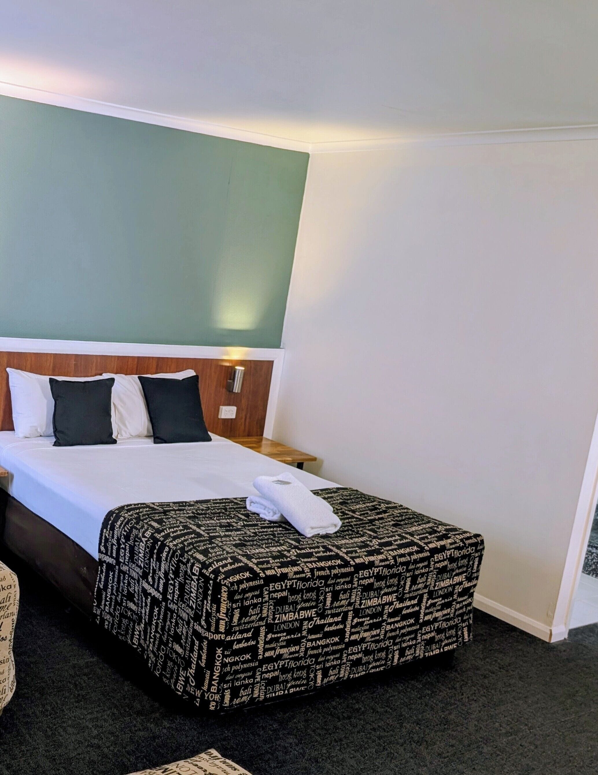 Una cama bien tendida con una colcha estampada, una mesita de noche con una lámpara y una pared verde decorativa.