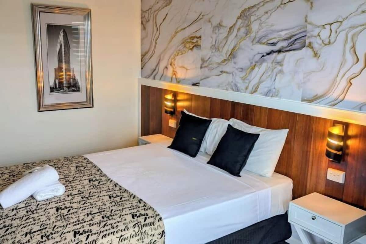 A hotel room with a bed, a nightstand, a wall-mounted light, and a framed picture on the wall.