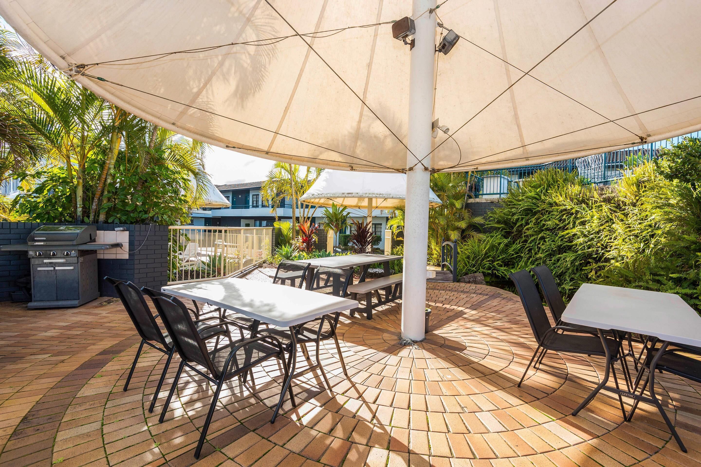 Une terrasse extérieure avec un sol en briques, des tables, des chaises et un grand parasol pour créer de l’ombre.