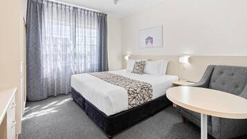 A hotel room with a bed, a small table, a grey armchair, and a window with curtains.