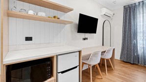 A modern kitchen with white cabinetry, a built-in dishwasher, a white refrigerator, a flat-screen TV mounted on the wall, and a dining area with two white chairs.