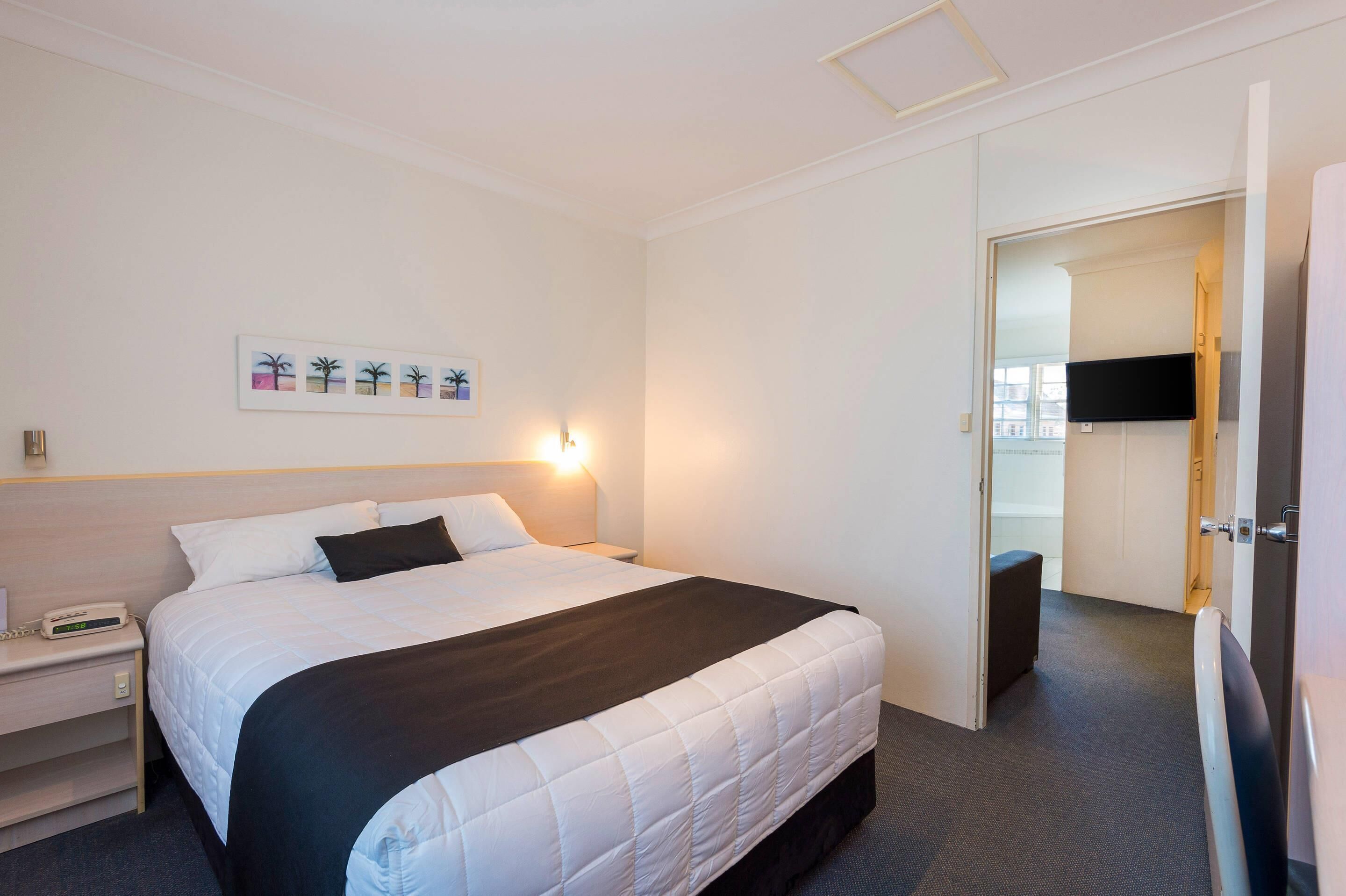 A hotel room with a bed, bedside table, lamp, artwork, and a television.