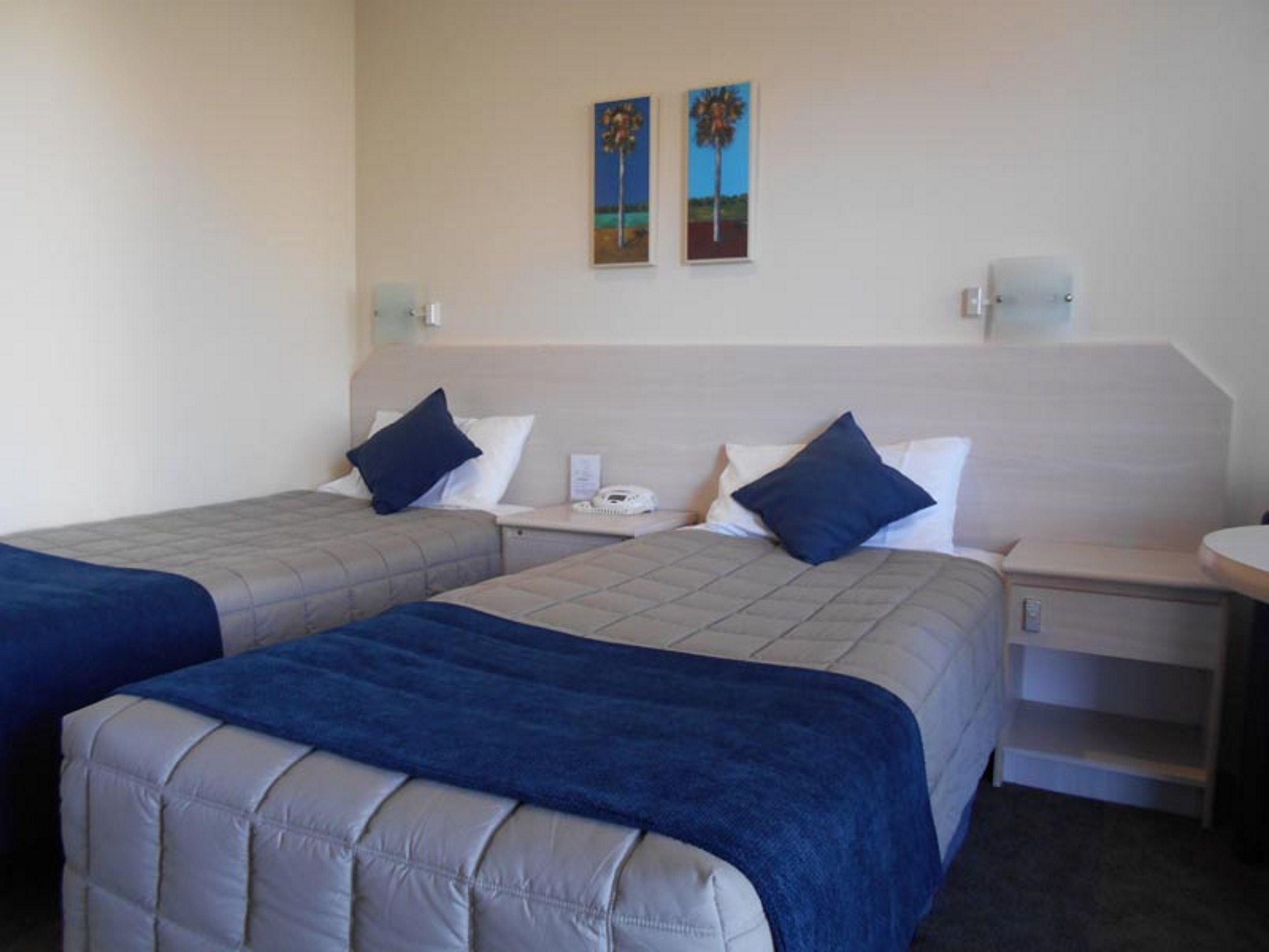 A hotel room with two beds, each with a blue bedspread and pillows, a bedside table with a phone, and a wall-mounted light.