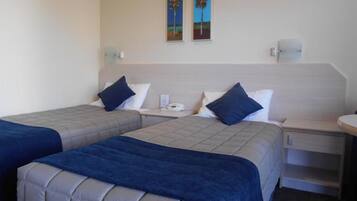 A hotel room with two beds, each with a blue bedspread and pillows, a bedside table with a phone, and a wall-mounted light.