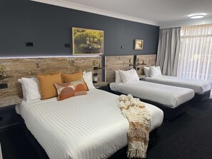 Two single beds with white linens and decorative pillows, complemented by a large framed picture and sheer curtains.