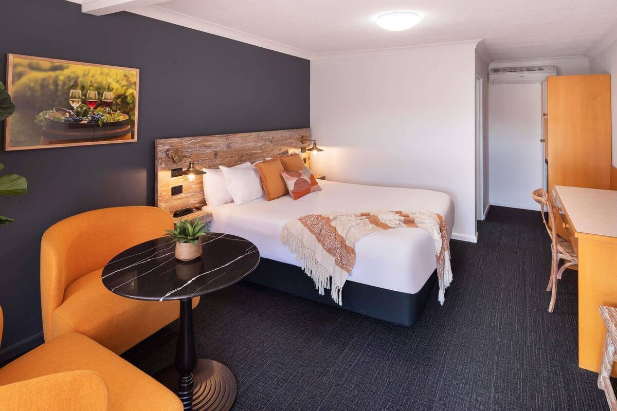 A hotel room with a bed, a small round table with a plant, an orange armchair, a wooden headboard, and a painting on the wall.