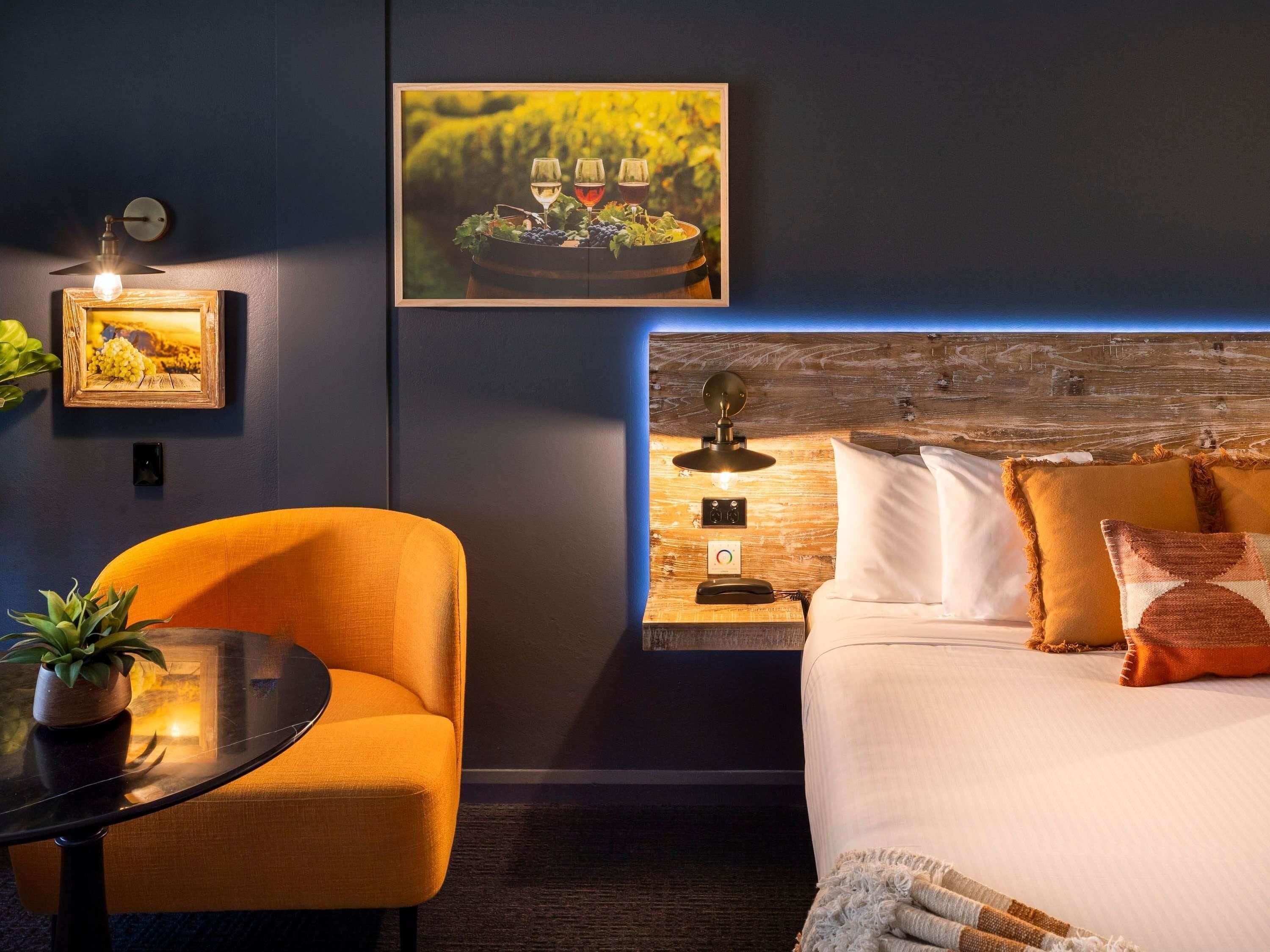 A modern hotel room with a bed, a nightstand, a table with a chair, and a wall-mounted picture of a vineyard.