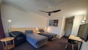 Quarto de hotel com duas camas de solteiro, uma escrivaninha com cadeira, um ventilador de teto e um quadro na parede.