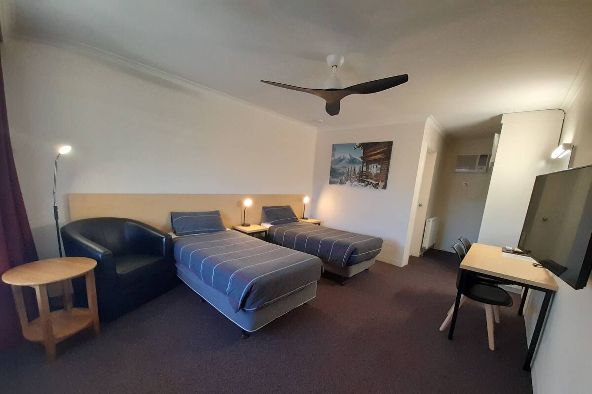 A hotel room with two single beds, a desk with a chair, a ceiling fan, and a painting on the wall.