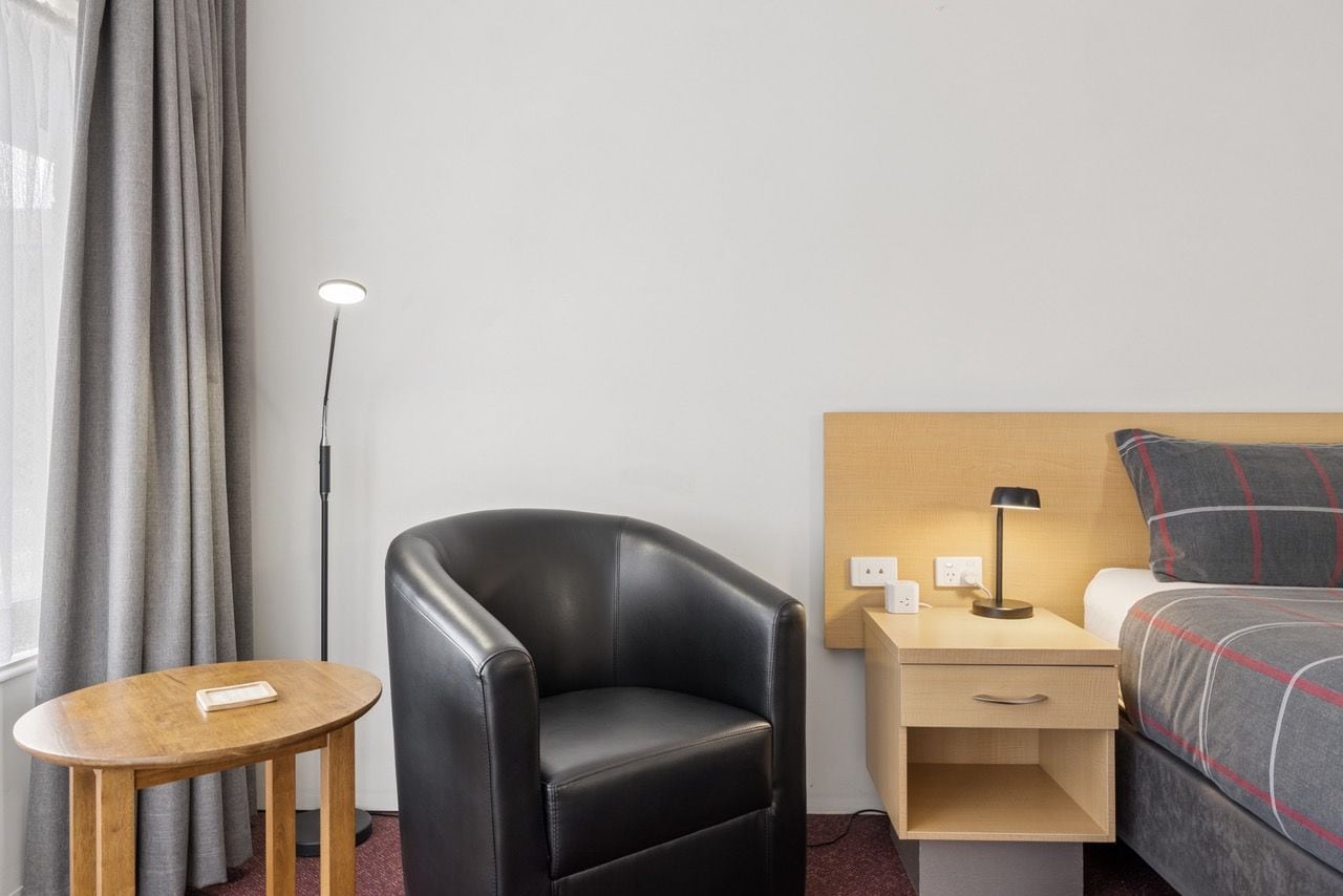 Ett hotellrum med en säng, ett skrivbord med en lampa, en stol, ett litet bord och en golvlampa.