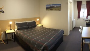 Quarto de hotel com cama, luminárias de cabeceira, escrivaninha com cadeira, cabideiro e janela com cortinas.