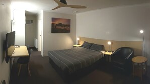 Quarto de hotel com cama, escrivaninha, cadeira, luminária e ventoinha no teto.
