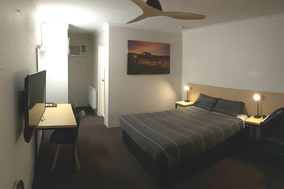 A hotel room with a bed, a desk, a chair, a lamp, and a ceiling fan.