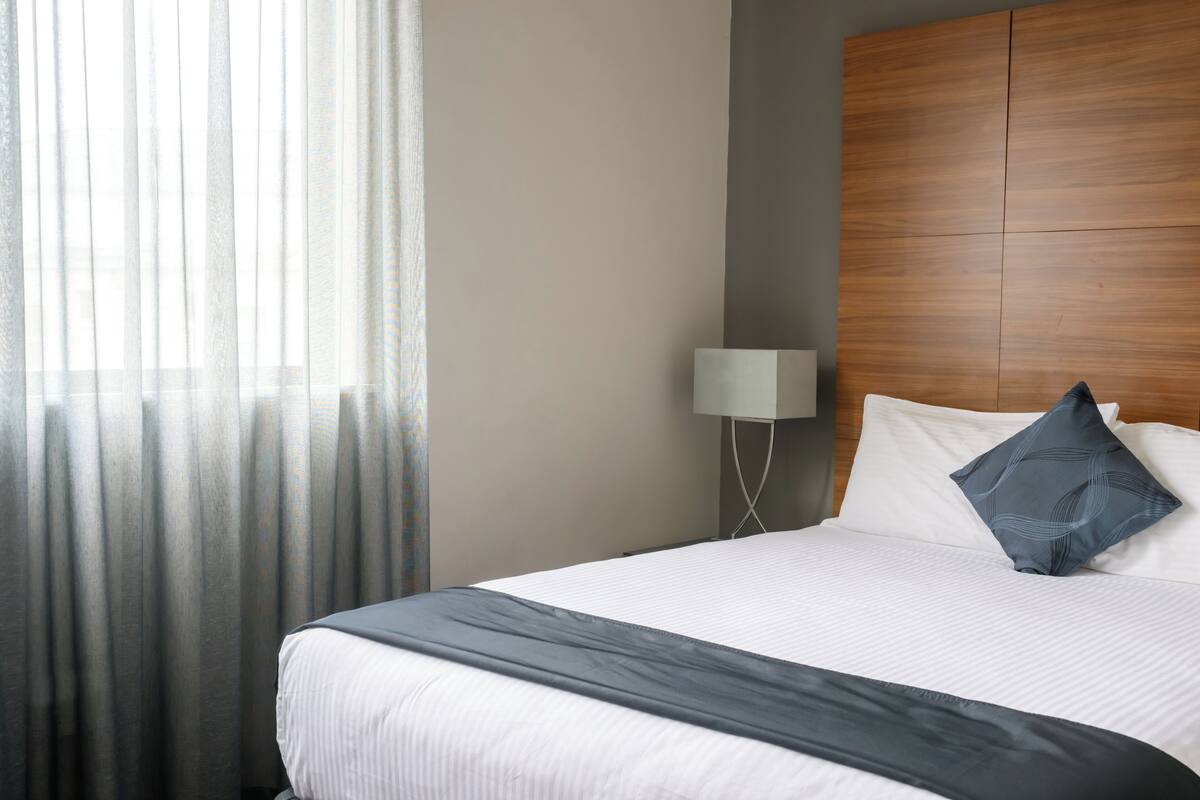 A hotel room with a bed, a nightstand, a lamp, and sheer curtains.