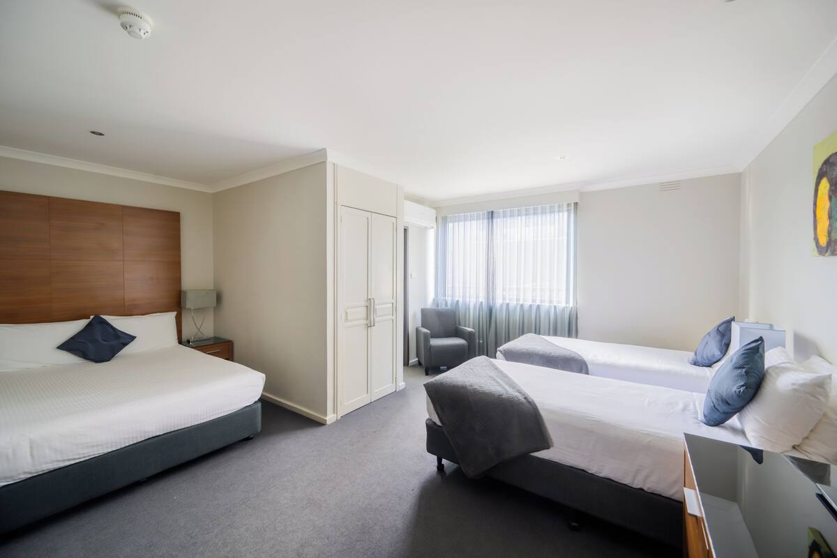 A hotel room with two single beds, a wooden headboard, a grey armchair, and a large window with curtains.