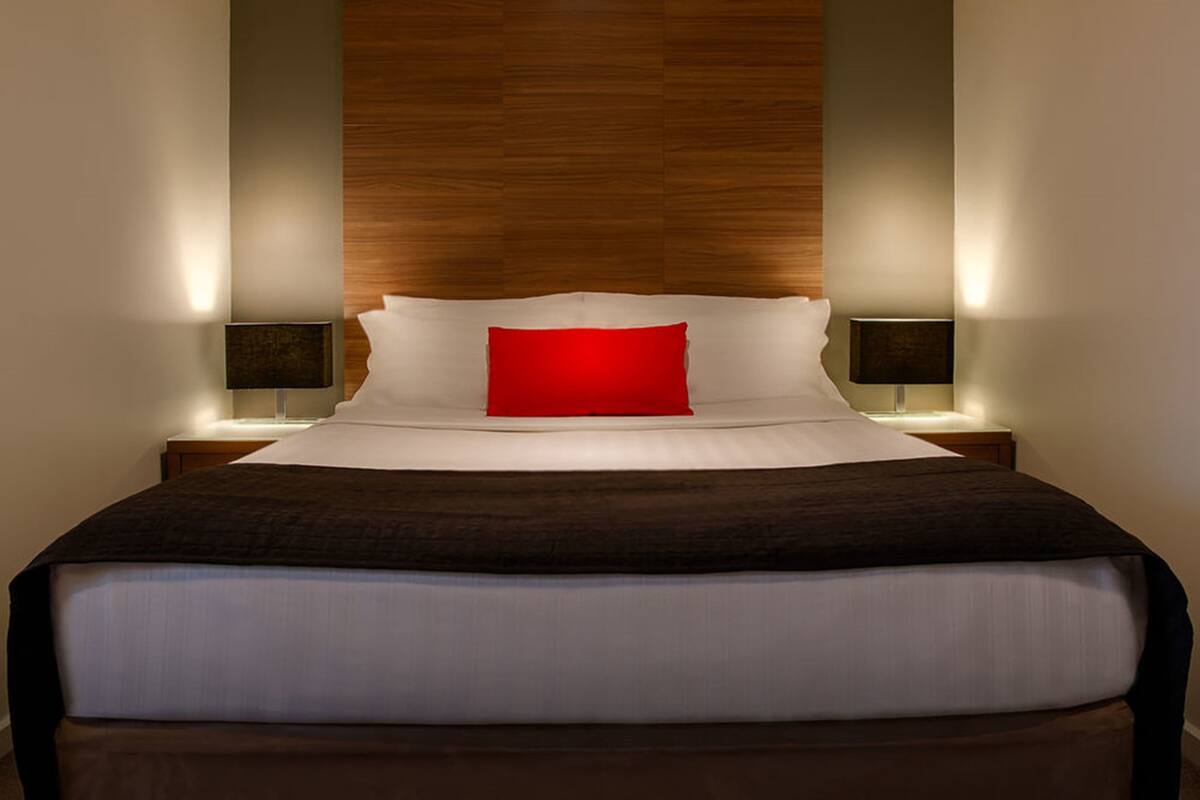 A neatly made bed with a red pillow, two bedside lamps, and a wooden headboard.