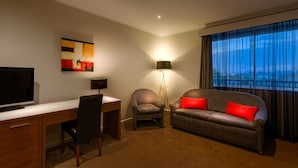 A hotel room with a desk, a sofa with red cushions, a chair, a floor lamp, and a painting on the wall. - Crest on Barkly (St Kilda)