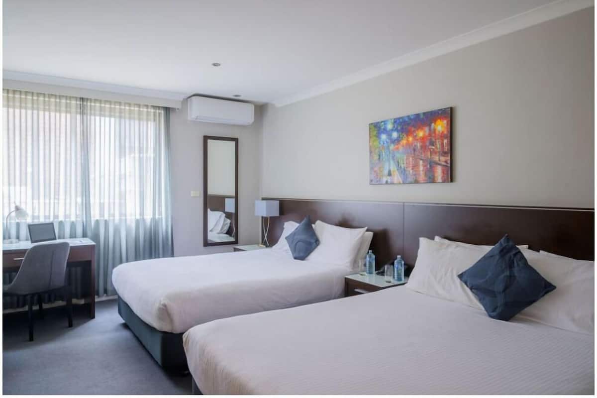 A hotel room with two beds, a desk, a chair, a mirror, a painting, and an air conditioning unit.