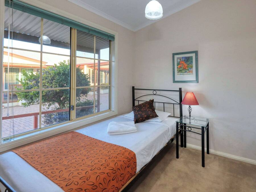 Un dormitorio con cama, ventana, mesita de noche con lámpara y un cuadro enmarcado en la pared.