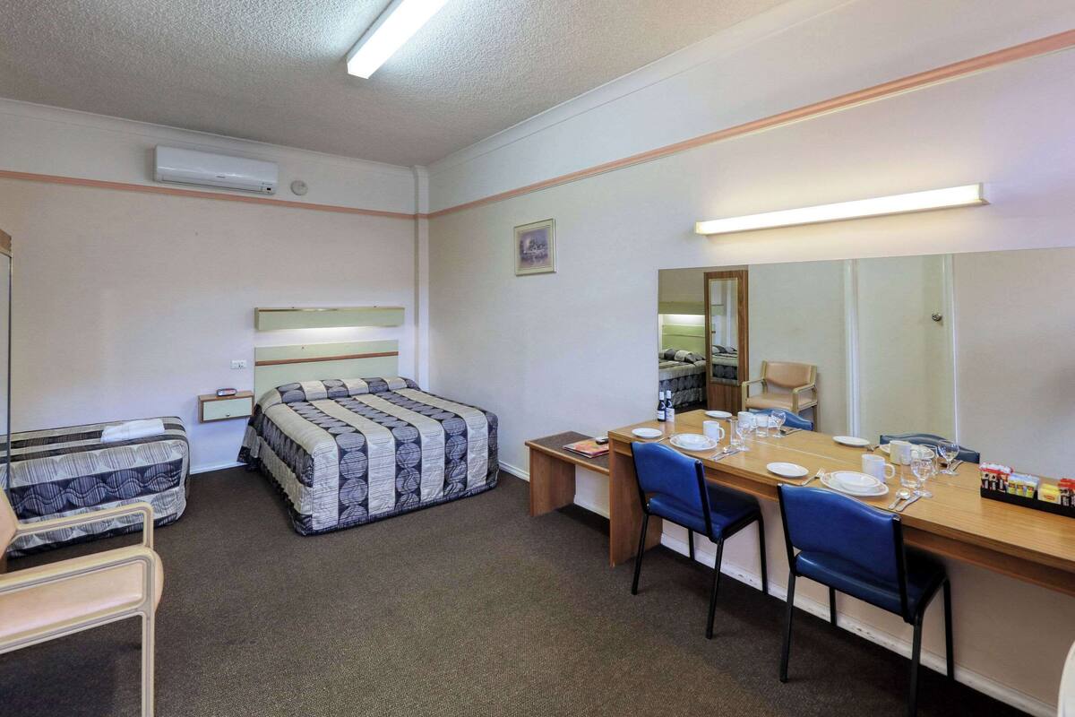 A hotel room with a bed, a desk with chairs, a mirror, and a small table with a lamp.