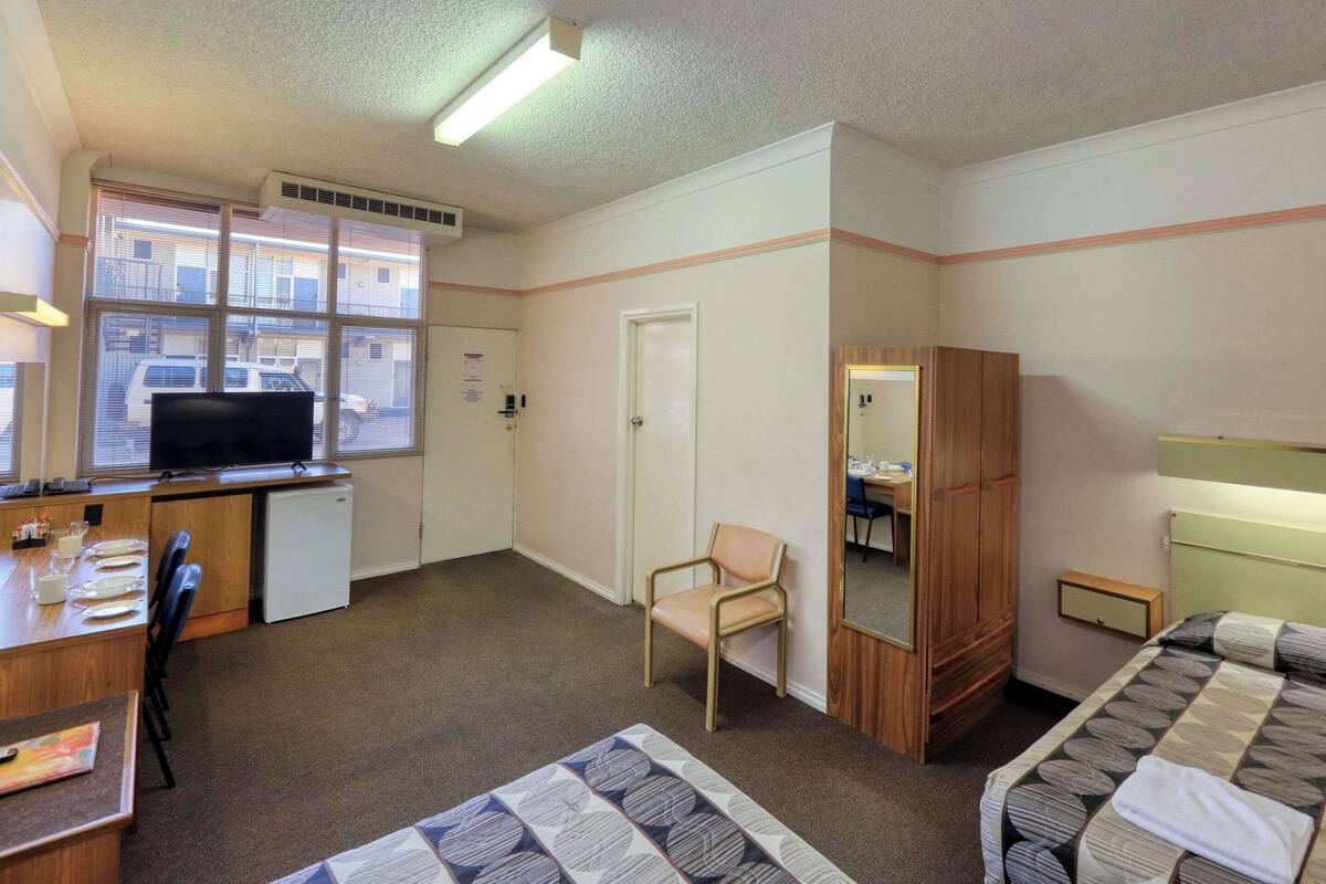 A hotel room with a bed, a desk with a computer, a chair, a wardrobe, and a window with blinds.