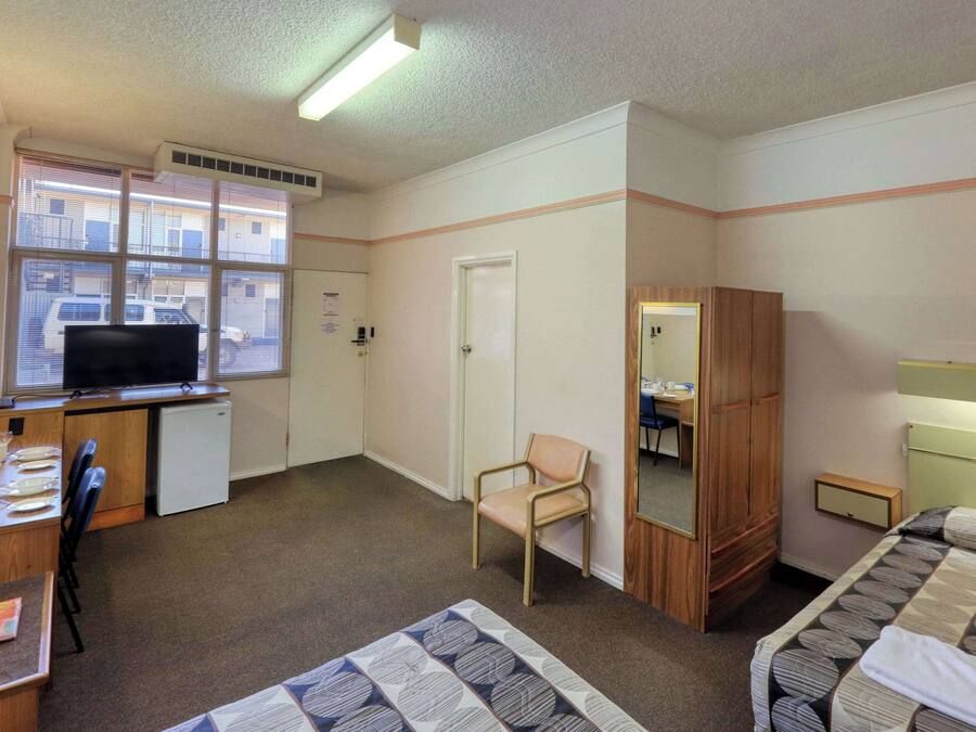 A hotel room with a bed, a desk with a computer, a chair, a wardrobe, and a window with blinds.