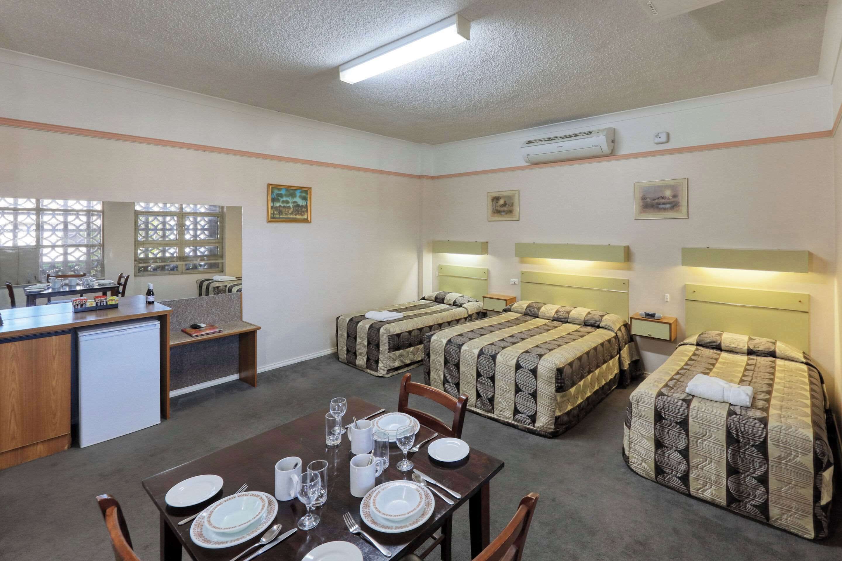 A compact hotel room with a kitchenette, two beds with headboards, a dining table set for four, and a small seating area.