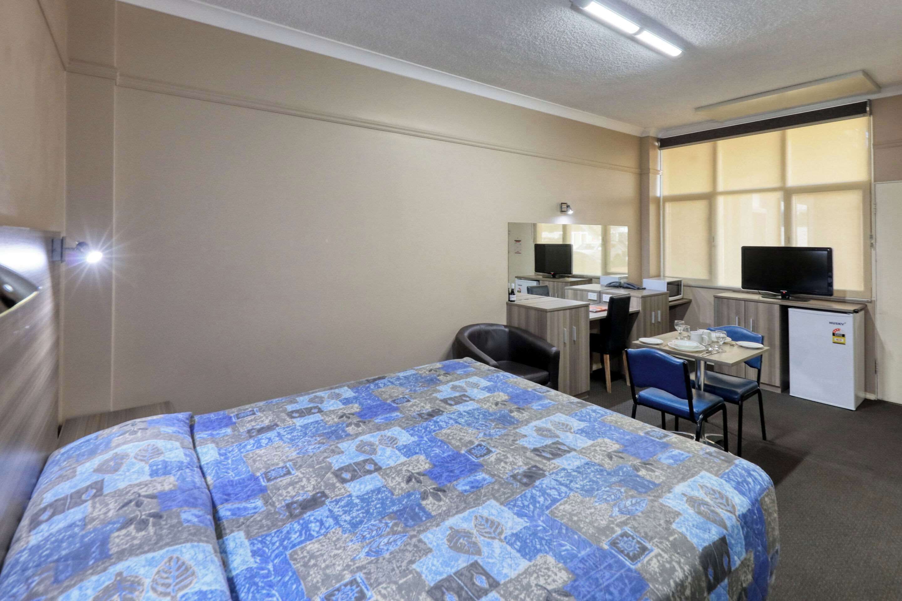 Una habitación de hotel con cama, escritorio, computadora, televisión y un refrigerador pequeño.