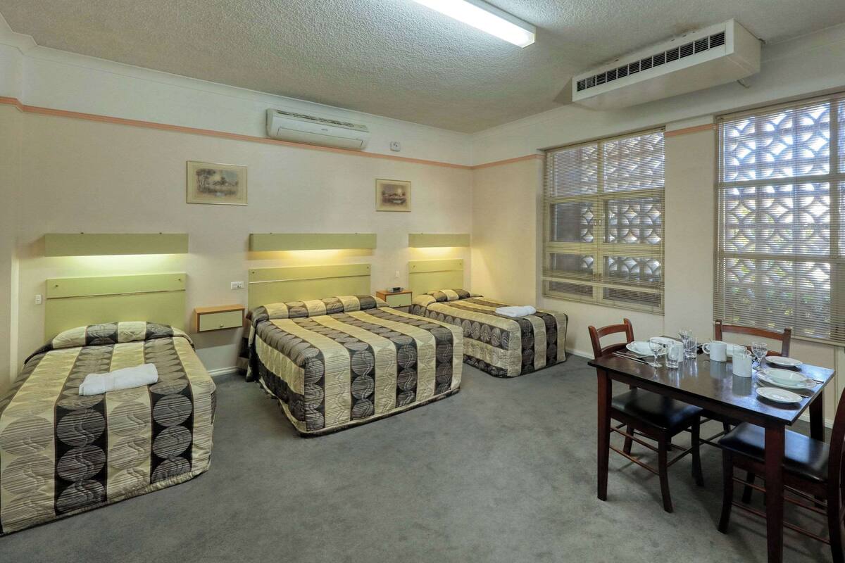 A room with three beds, a dining table with chairs, and a window with blinds.