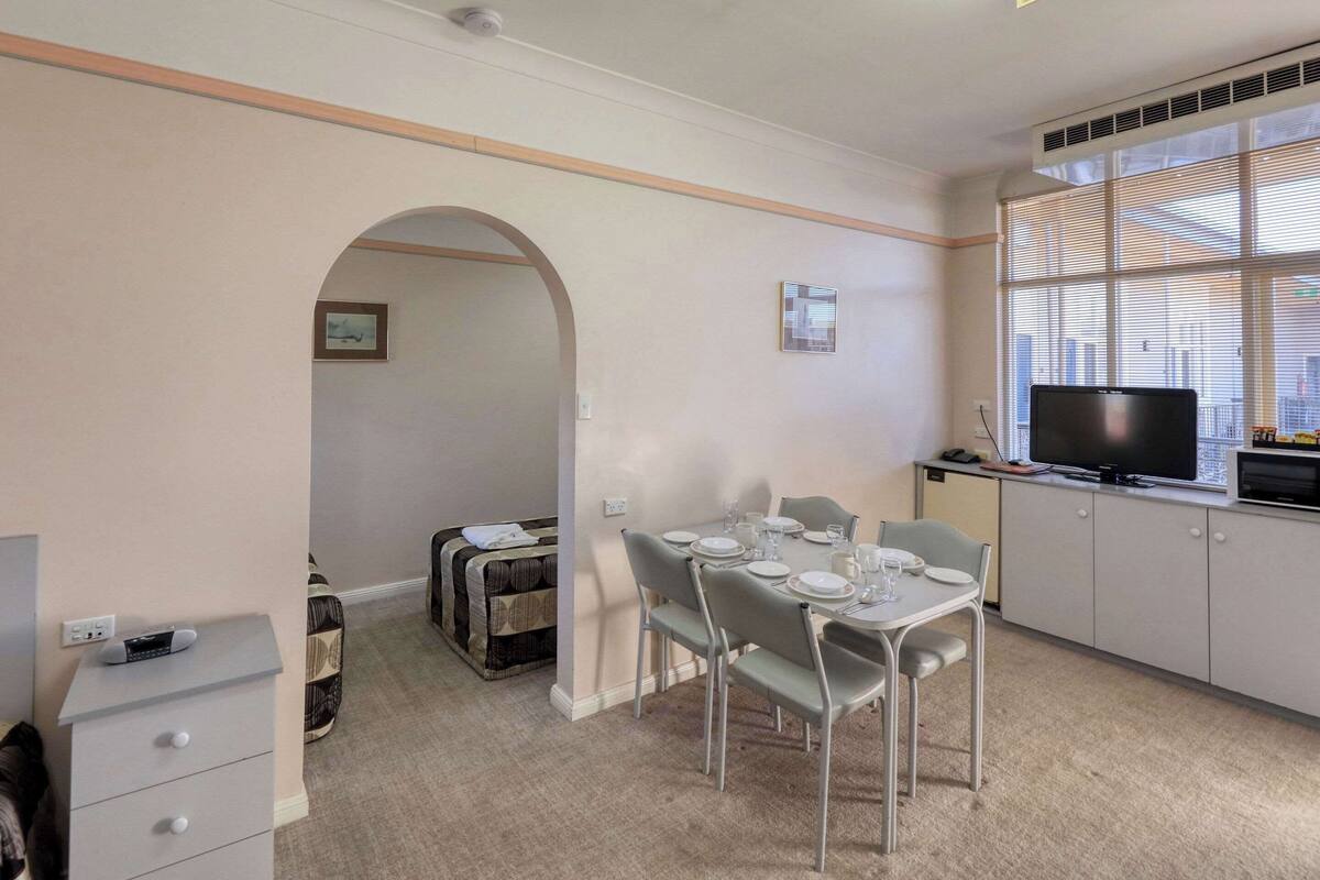 A dining area with a table set for four, a television on a cabinet, and a window with blinds.