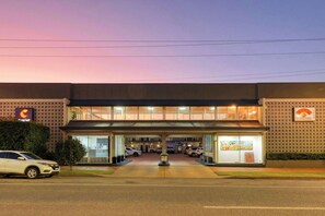 Un edificio moderno con acceso cubierto, varios espacios de estacionamiento y un letrero visible a la izquierda.