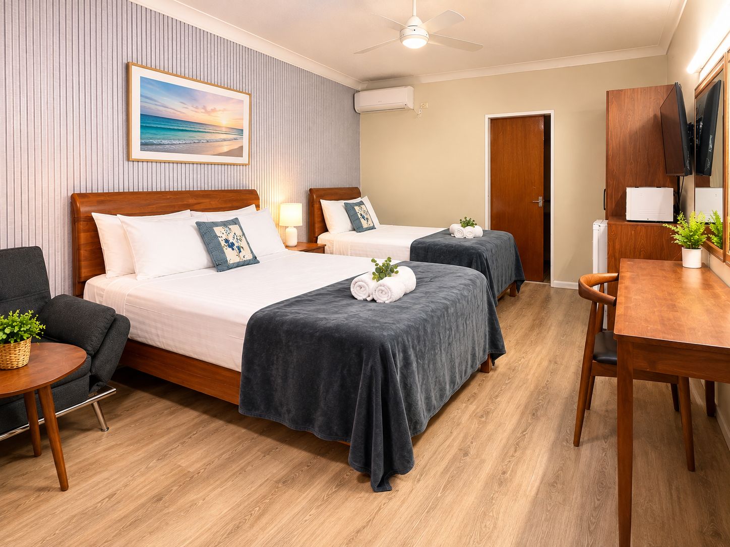 A hotel room with two beds, a desk, a chair, a small table with a plant, and a ceiling fan.