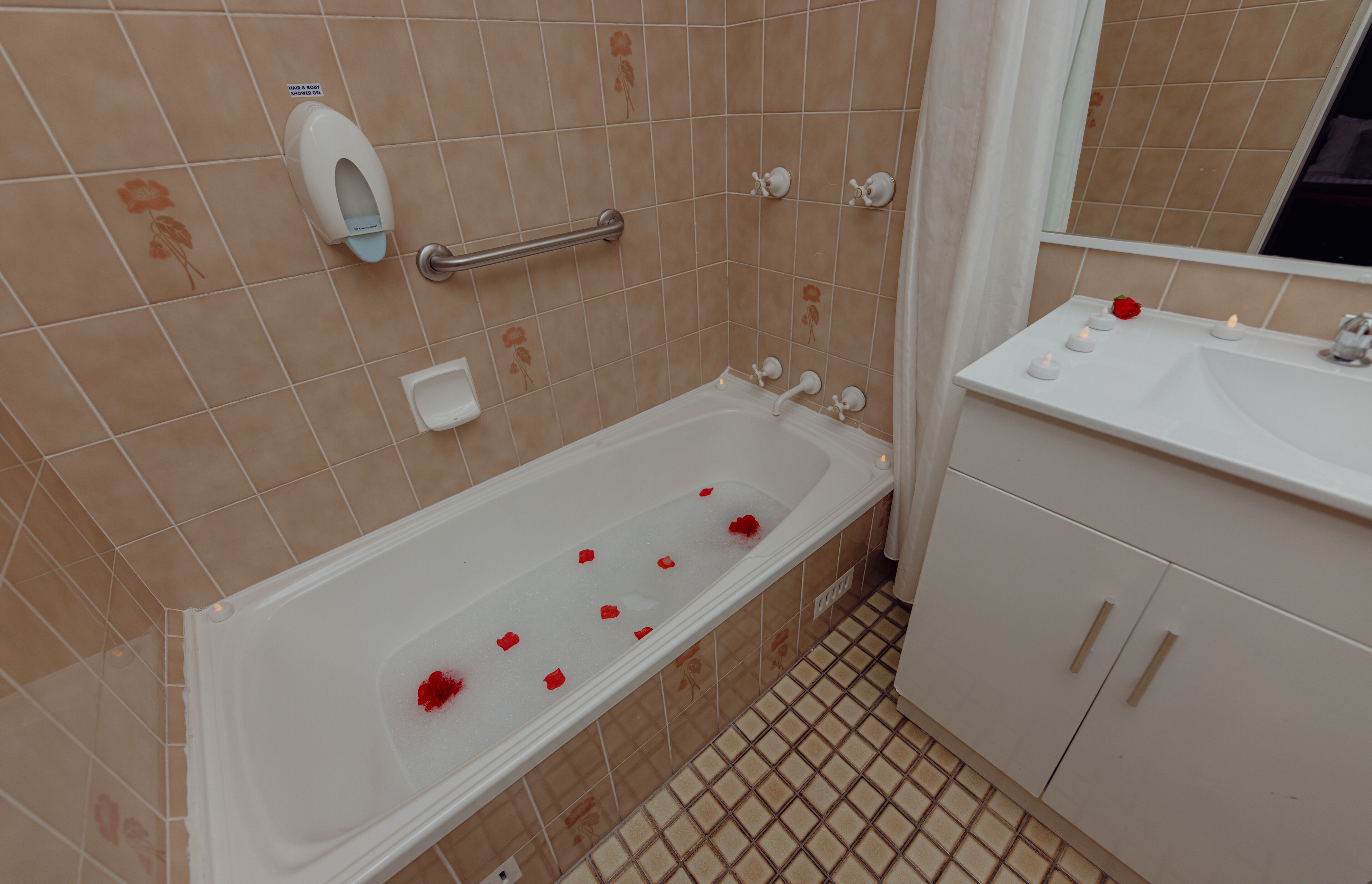 A bathroom with a bathtub, a hand dryer, a hand soap dispenser, a hand rail, a sink, and a mirror.