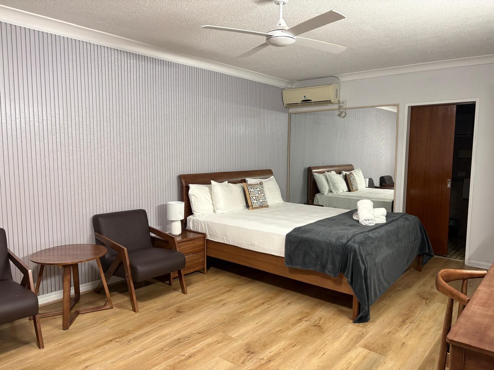A hotel room with a bed, bedside tables, a chair, a desk, a ceiling fan, and an air conditioning unit.