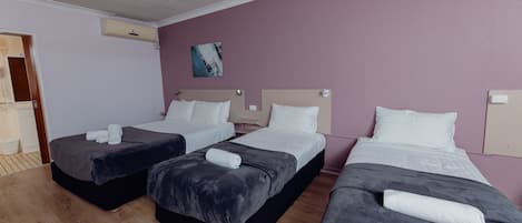 Habitación de hotel con tres camas, cada una con una colcha oscura y almohadas blancas, y un ventilador de techo.