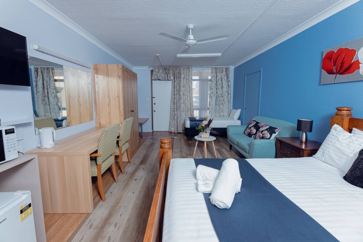 A hotel room with a bed, a desk, a chair, a TV, a ceiling fan, and a window with curtains.