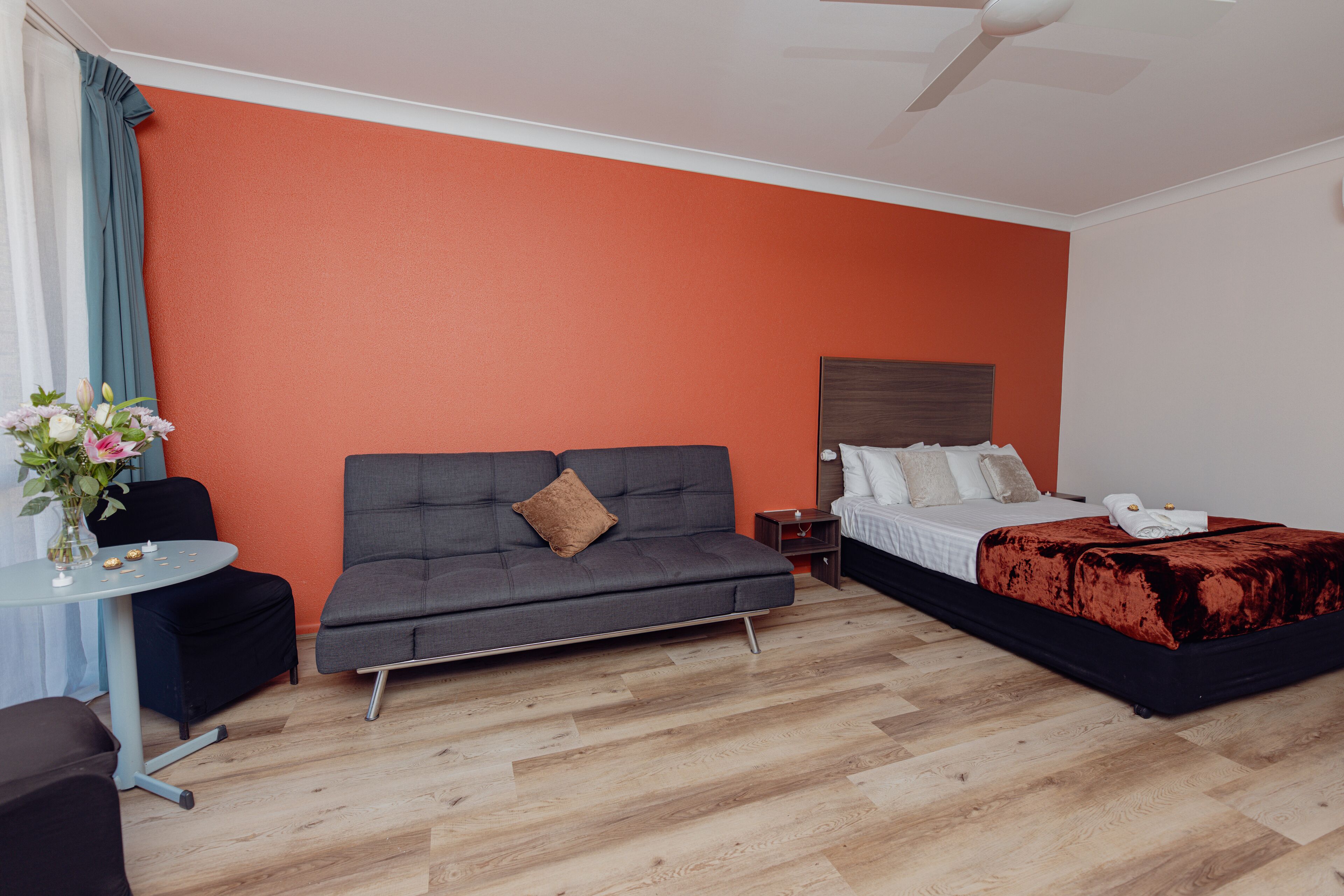 A hotel room with a grey sofa, a bed with a red blanket, a small table with a vase of flowers, and a ceiling fan.