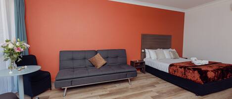 A hotel room with a grey sofa, a bed with a red blanket, a small table with a vase of flowers, and a ceiling fan.