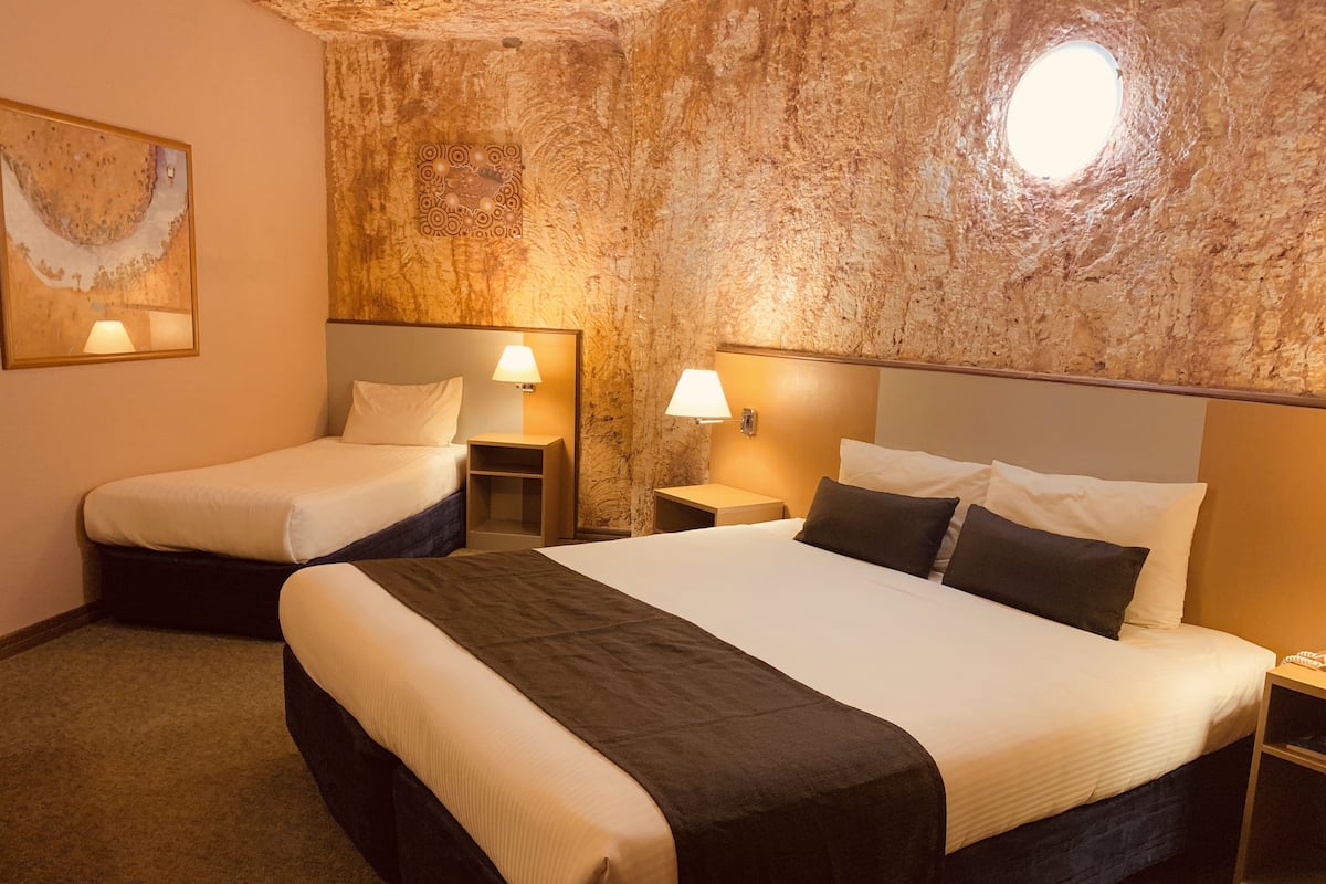 A hotel room with two single beds and one double bed, each with a bedside table and lamp. The room features a textured stone wall and a painting on the left.