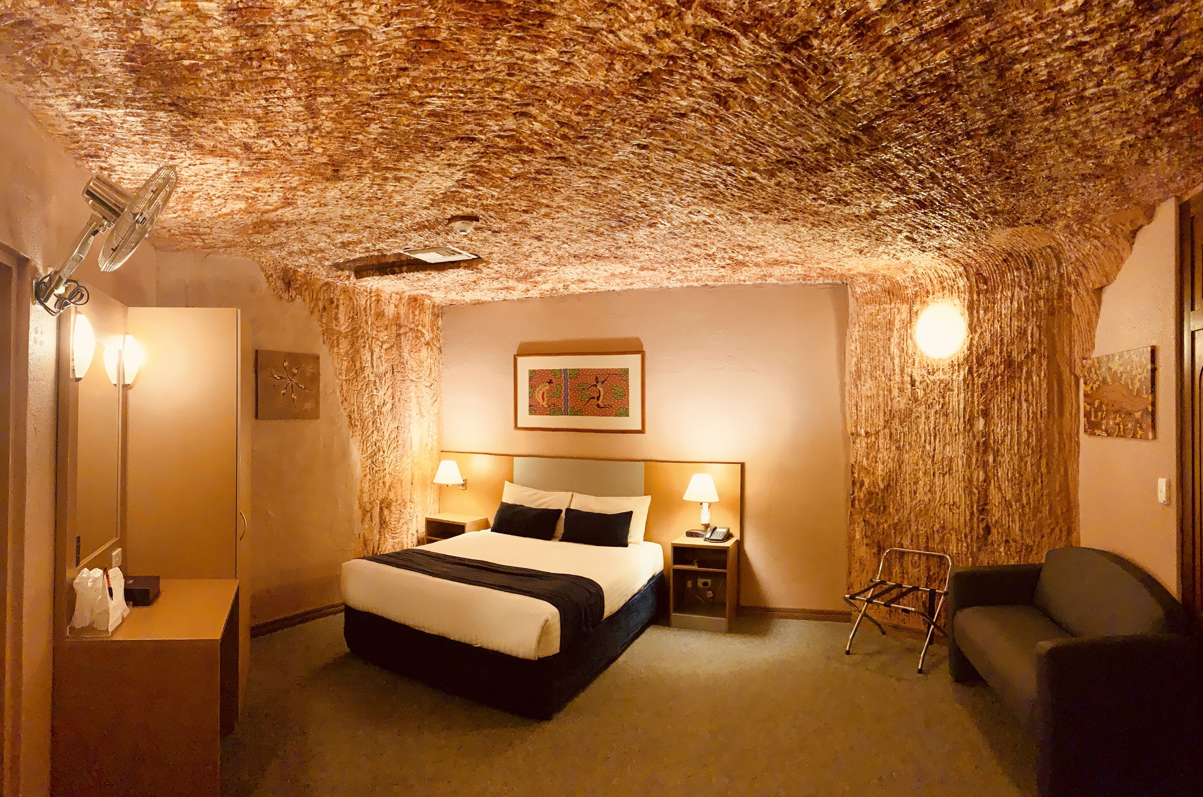 A hotel room with a unique textured ceiling, a bed with a black and white bedspread, two bedside lamps, a nightstand with a phone, a chair, and a small table with a lamp.