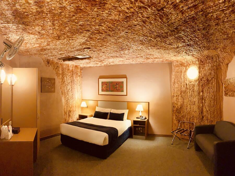A hotel room with a unique textured ceiling, a bed with a black and white bedspread, two bedside lamps, a nightstand with a phone, a chair, and a small table with a lamp.