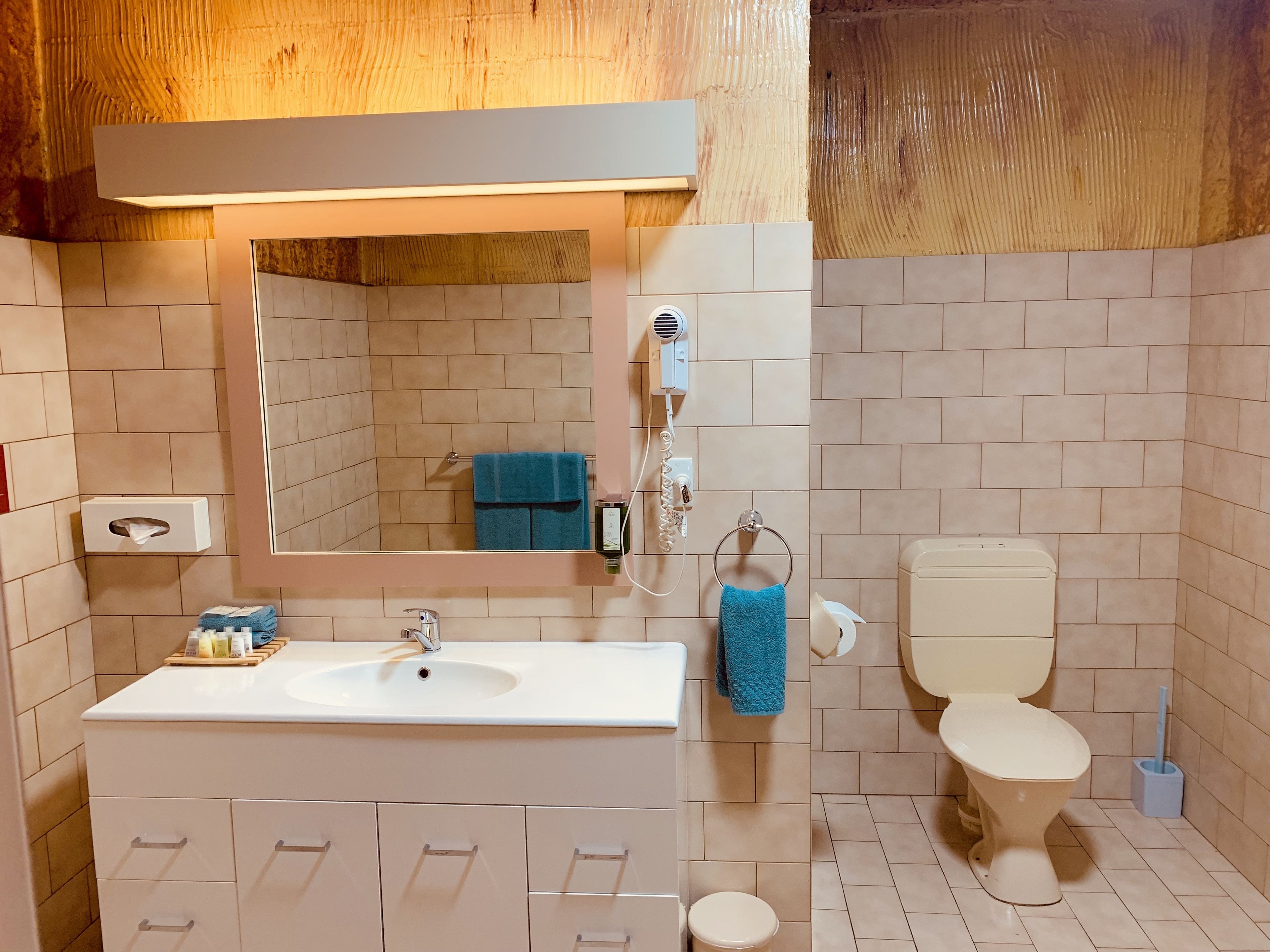 Een badkamer met een wit wastafel, spiegel en toilet. Er is een handdoekhouder met een blauwe handdoek, een zakdoekendoos en een haardroger. De muren zijn belegd met lichtbeige tegels.