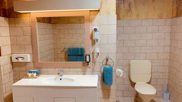 Een badkamer met een wit wastafel, spiegel en toilet. Er is een handdoekhouder met een blauwe handdoek, een zakdoekendoos en een haardroger. De muren zijn belegd met lichtbeige tegels.